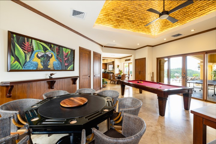 Room with poker and pool tables, artwork of macaws, and a view of outdoors through large windows.