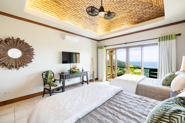 Bright bedroom with two beds, large window with ocean view, decorative mirror, TV, and ceiling fan.