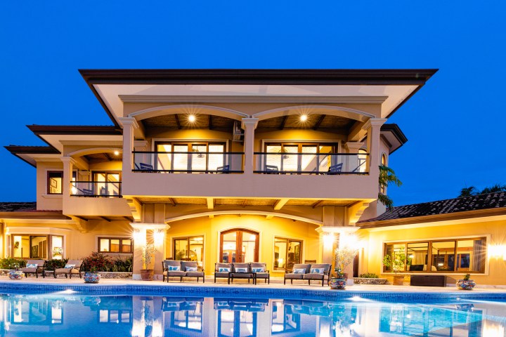 Illuminated two-story house with pool at dusk, featuring large windows and balconies.
