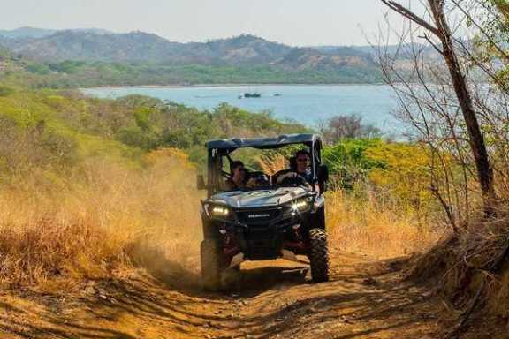Two people ride an ATV on a dirt trail near a lake surrounded by hills and sparse vegetation.