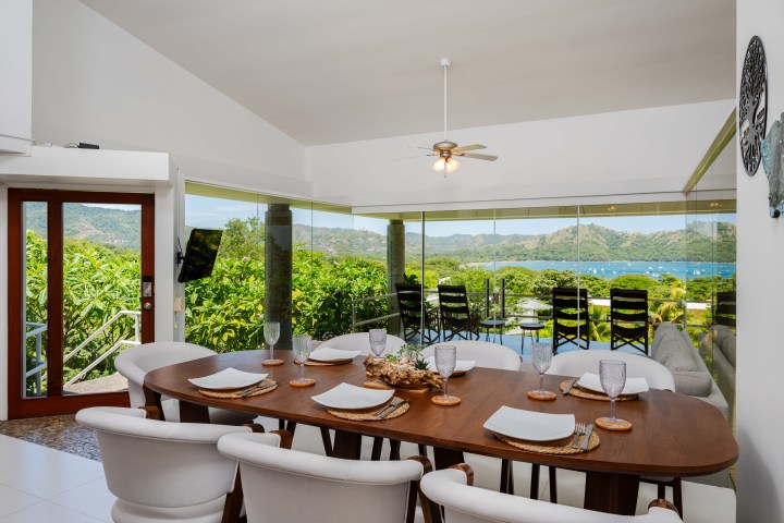 Dining room with a large wooden table, chairs, and a view of greenery and a bay through floor-to-ceiling windows.