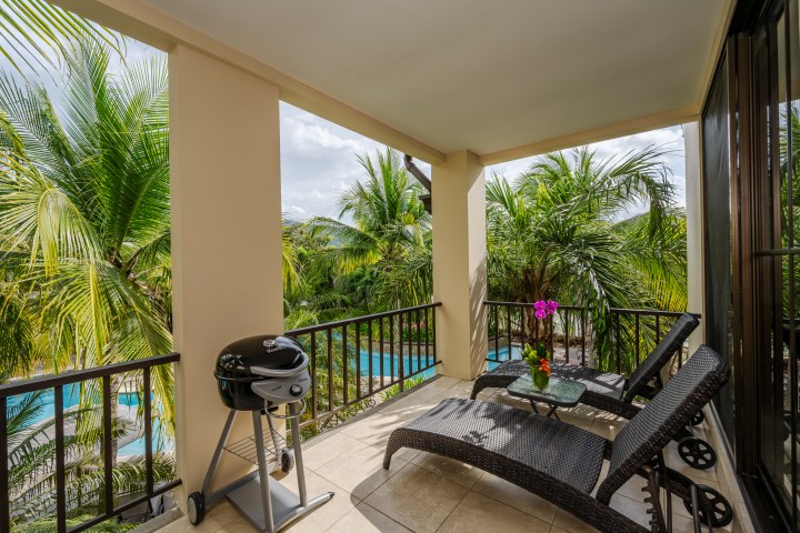 Balcony with two lounge chairs, table with flowers, and a grill overlooking palm trees and pool.