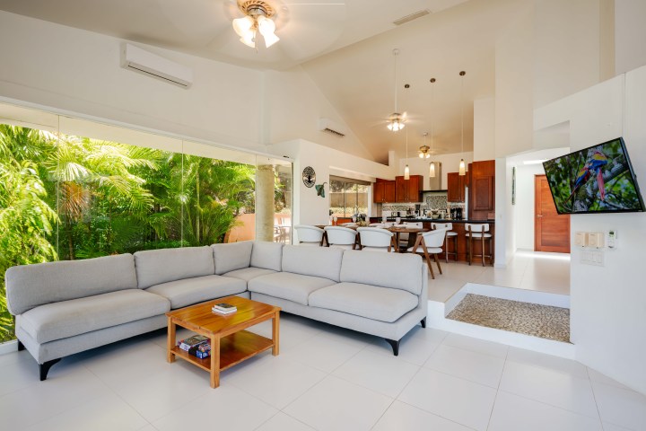 Modern living room with gray sectional sofa, wooden table, and open kitchen, large windows showing greenery.