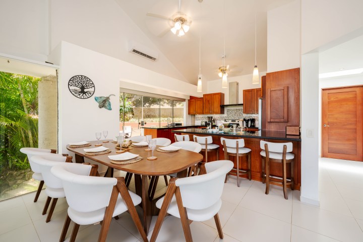 Modern kitchen and dining area with wooden furniture and white chairs, featuring pendant lighting.
