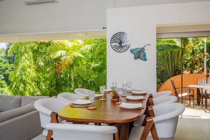 Dining area with wooden table, white chairs, large windows, and tropical view.