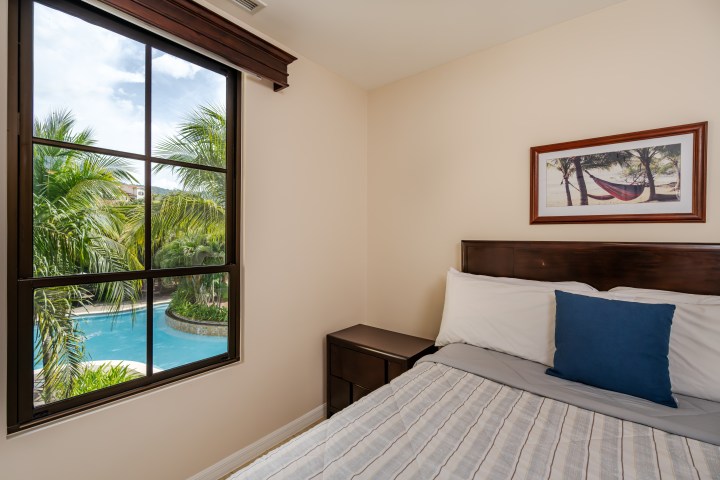Bedroom with bed, window showing palm trees and pool, framed beach photo on wall.