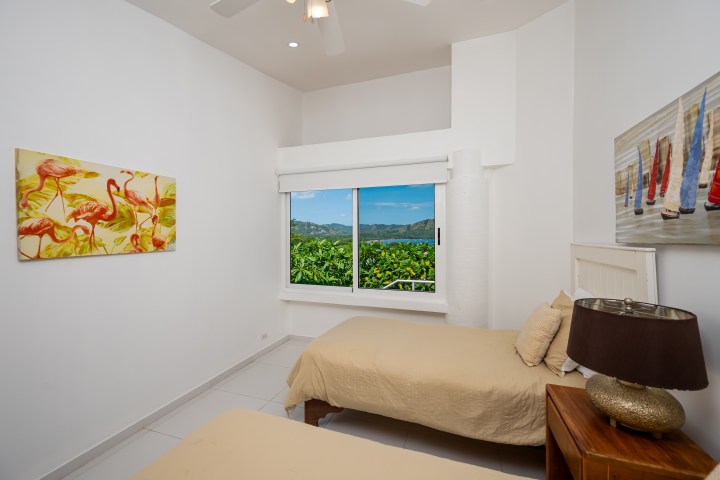 Room with two beds, tropical paintings, and a window view of hills.