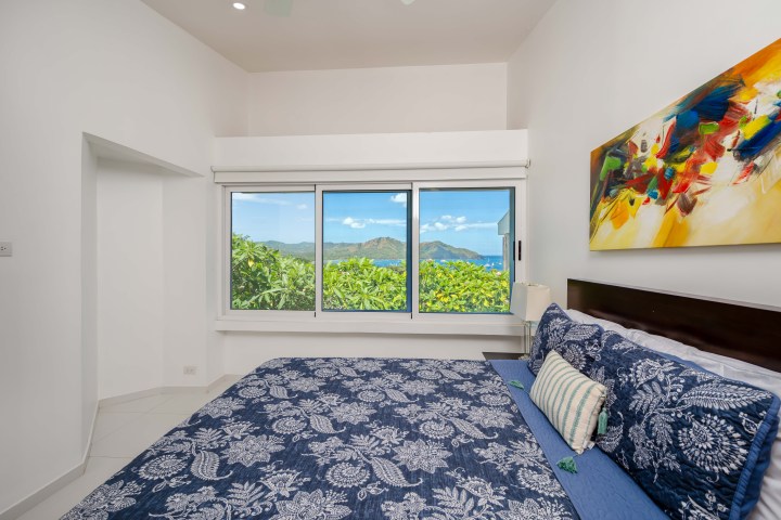 Bedroom with blue floral bedding, colorful wall art, and window view of ocean and greenery.
