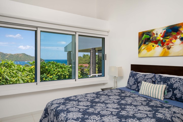 Bedroom with patterned bedding, window view of ocean and trees, abstract painting on wall.