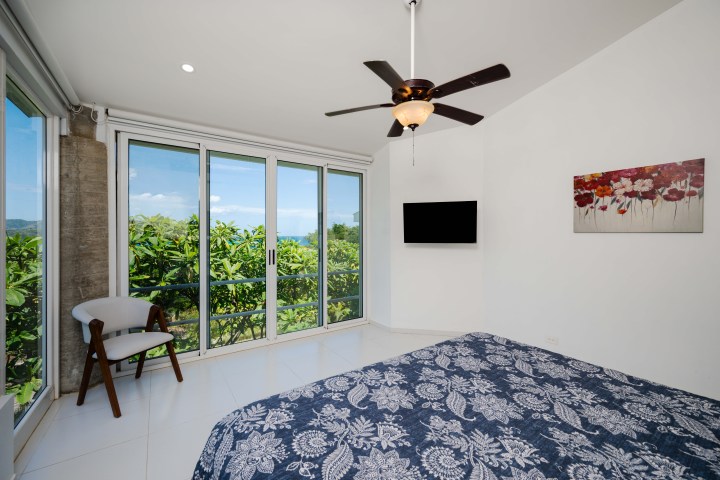 Bedroom with large windows, ceiling fan, bed, chair, TV, and floral artwork.
