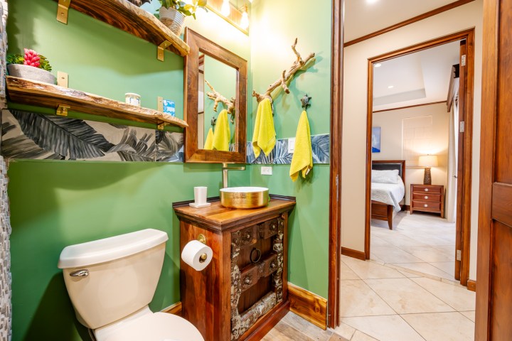 Stylish bathroom with green walls, wooden shelves, round mirror, yellow towels, and a glimpse of a bedroom.