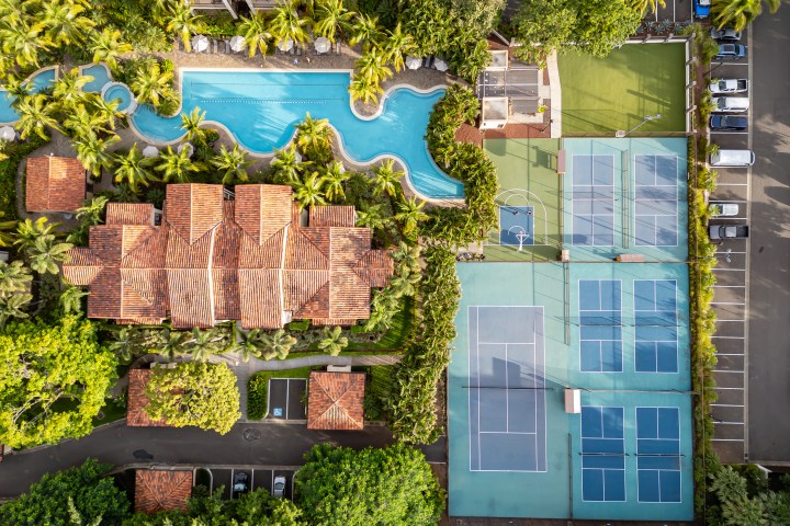 Aerial view of a resort with pool, tennis courts, and parking lot surrounded by greenery.