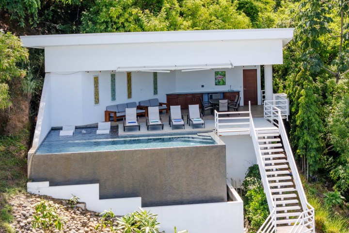 Modern hillside villa with pool, lounge chairs, bar area, and surrounded by lush greenery.