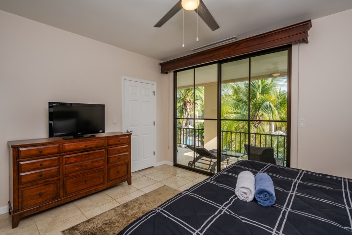 Bedroom with TV, dresser, bed, and large glass doors opening to a balcony with palm trees view.