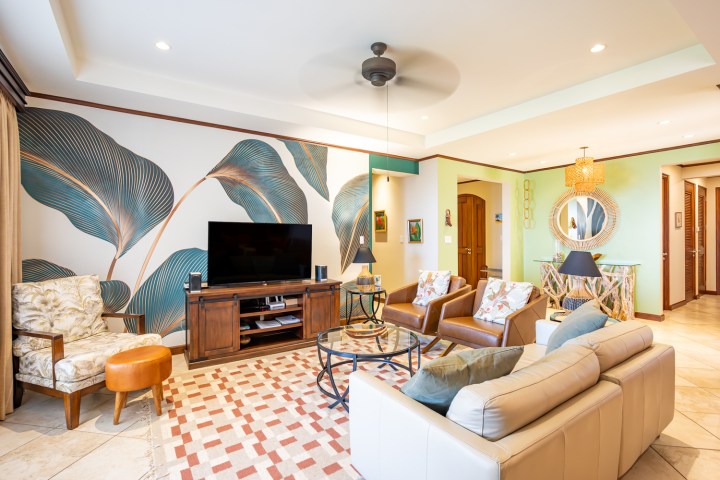 Living room with floral wallpaper, TV, beige sofa, armchairs, and decorative mirror on green wall.