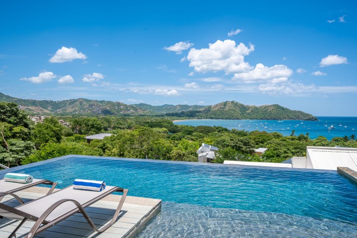 Infinity pool with lounge chairs overlooking a tropical bay and hills under a clear blue sky.