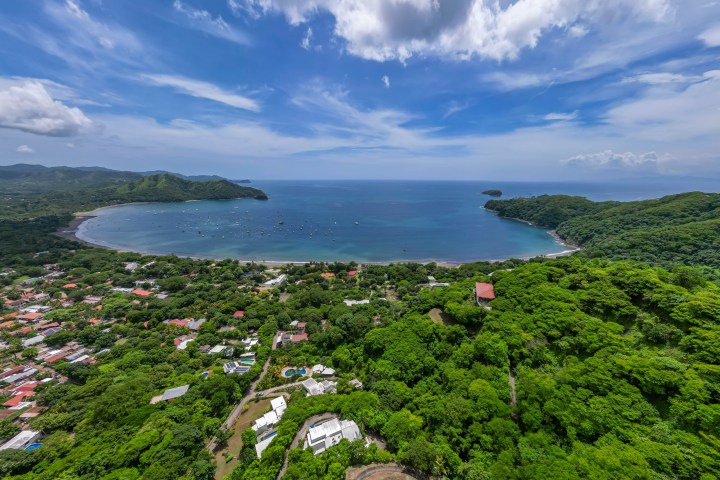 Aerial view of a lush coastal town with a bay, green hills, and blue sky.