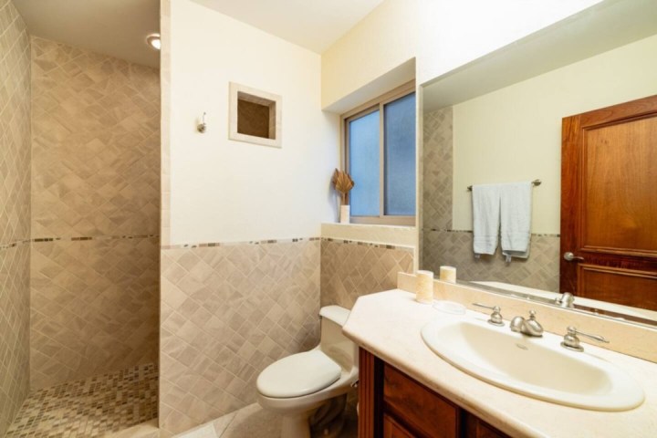 Bathroom with tiled walls, shower, toilet, sink, mirror, towel rack, and wooden door.