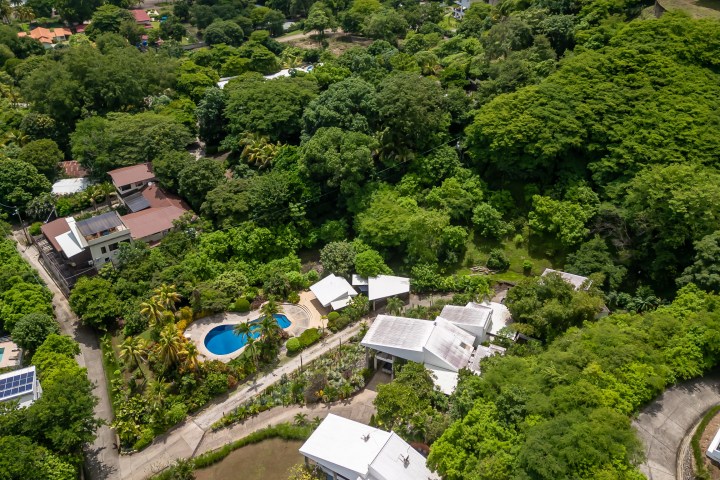 Aerial view of coastal area with houses, a pool, lush greenery, and a blue ocean in the background.