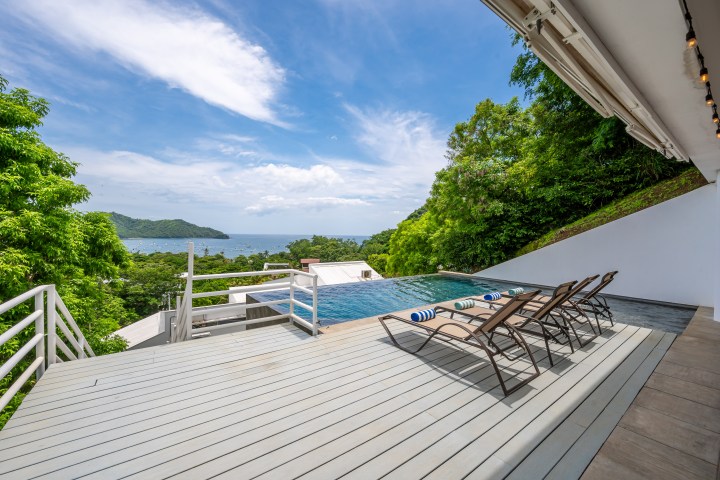 Poolside deck with lounge chairs overlooking ocean and green hills under a blue sky.