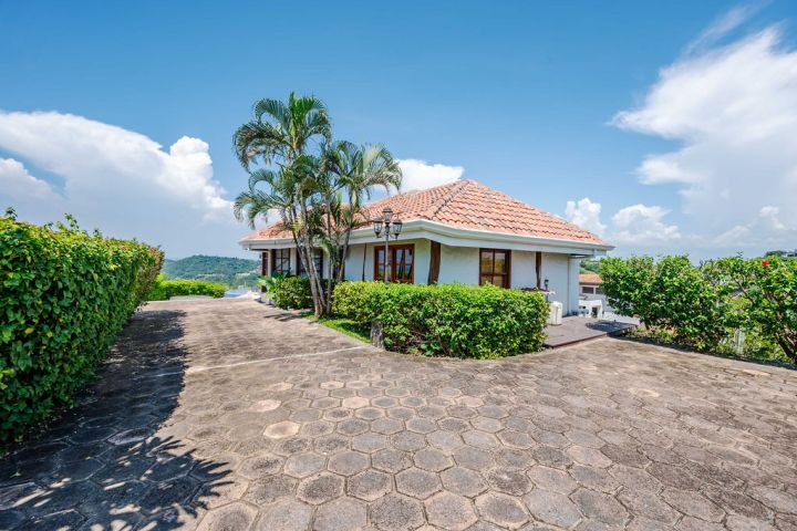 Single-story house with a red-tiled roof and palm trees, surrounded by greenery under a clear blue sky.