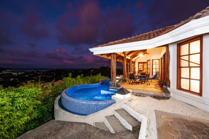 Night view of a patio with infinity pool and seating area overlooking a hilly landscape.