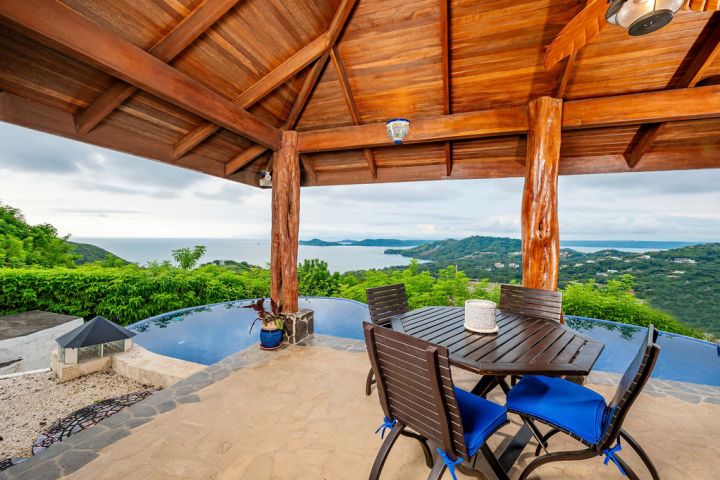 Outdoor patio with wooden roof, table, and chairs overlooking ocean and hills.