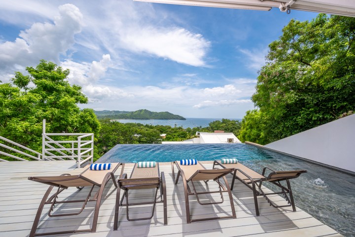 Four lounge chairs by an infinity pool overlooking a lush landscape and distant ocean views.