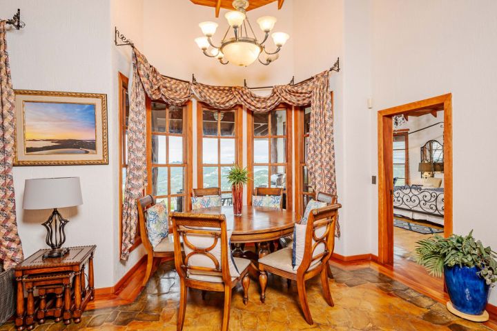 Dining room with round table, wooden chairs, chandelier, and large windows with patterned curtains.