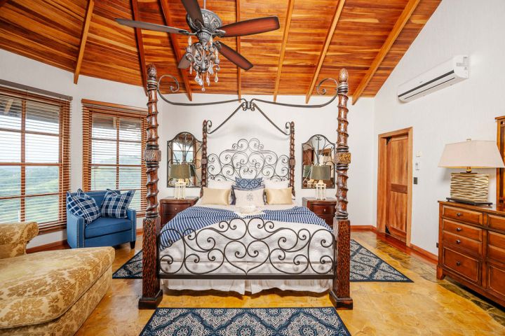Elegant bedroom with wooden ceiling, ornate bed, blue armchair, and views through large windows.