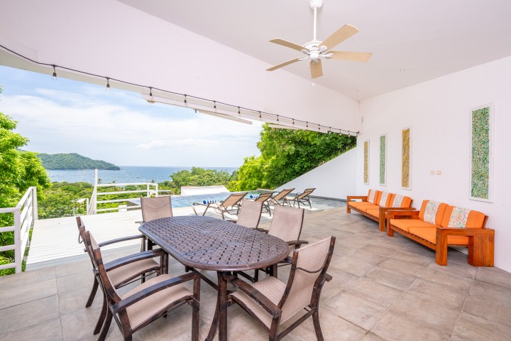 Open patio with dining table, orange couch, ceiling fan, view of pool, trees, and ocean.