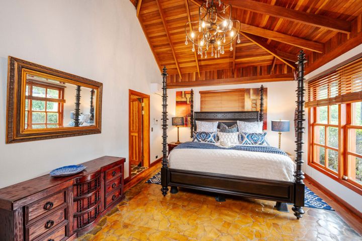 Bedroom with canopy bed, ornate mirror, wooden ceiling, and large windows with blinds.