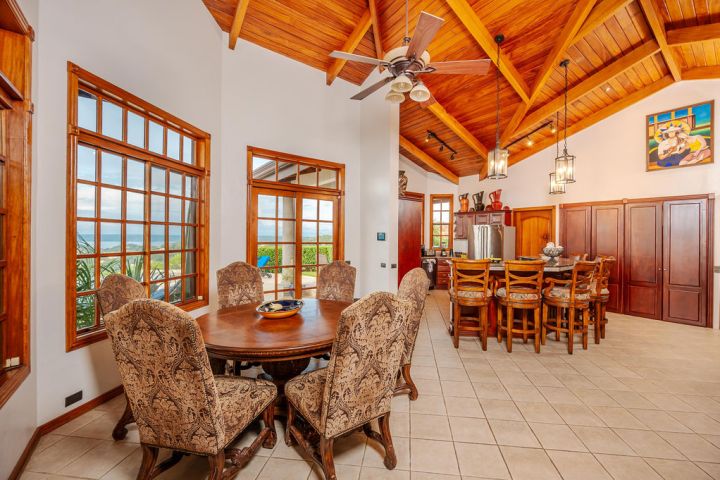 Dining area with round table, upholstered chairs, wooden ceiling, bar stools, and large windows.