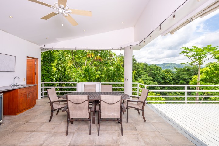 Outdoor patio with table, chairs, ceiling fan, and distant view of green trees and mountains.