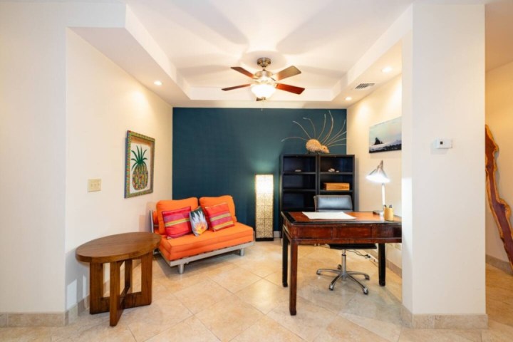 Modern office with orange sofa, wooden desk, ceiling fan, artwork, and shelving in a beige room.
