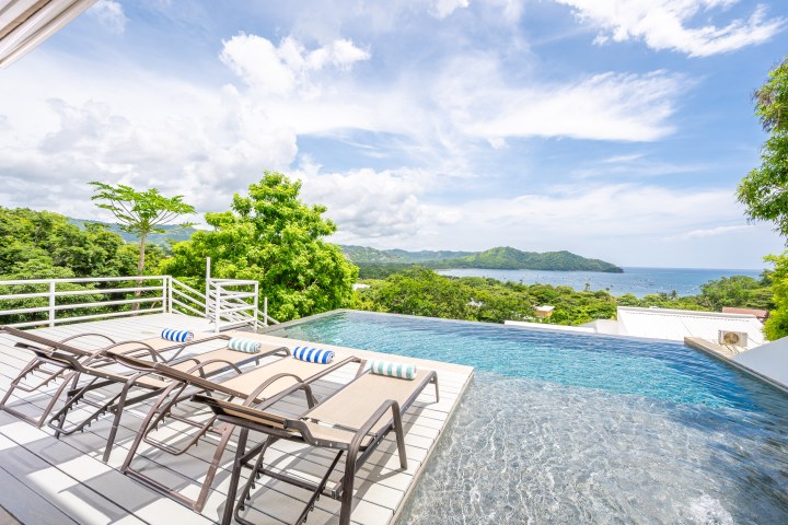 Infinity pool with lounge chairs overlooking ocean and hills on a sunny day.