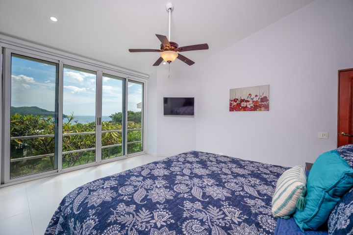 Bedroom with sea view, bed, ceiling fan, TV, and floral artwork.