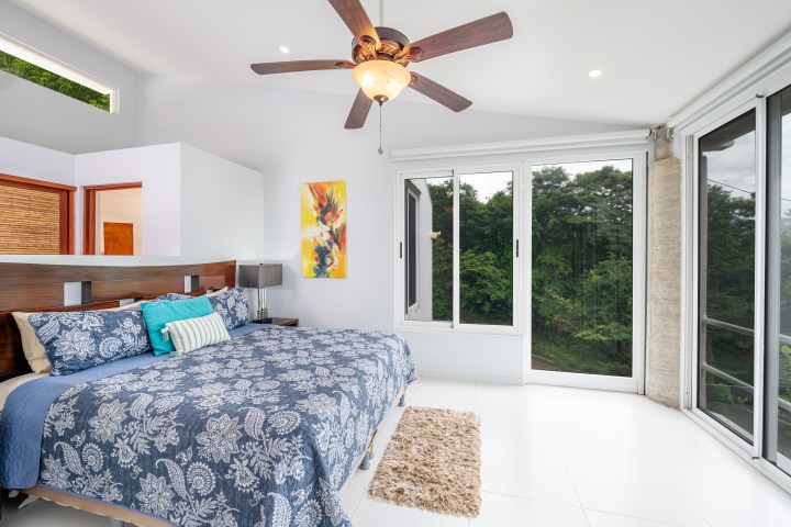 Bright bedroom with a ceiling fan, blue bedding, abstract painting, and large windows with a forest view.