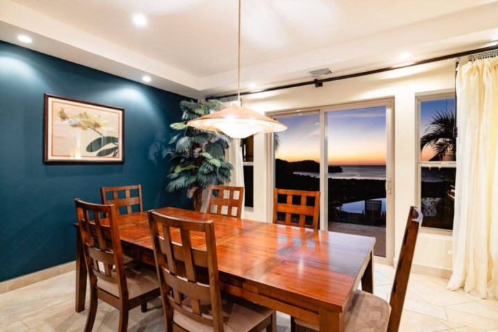Dining room with wooden table, six chairs, and a sunset view through large windows.
