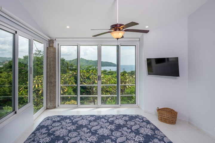 Bedroom with large windows, ocean view, ceiling fan, TV, and blue bedspread.