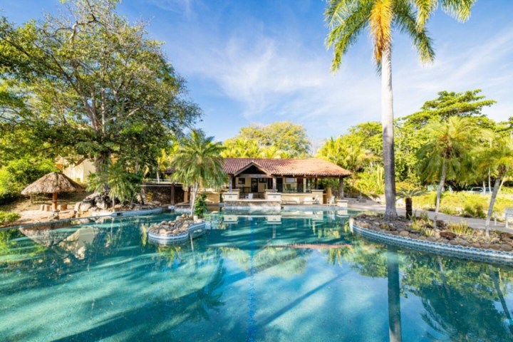Pool surrounded by tropical trees, with a thatched hut and building in the background under a clear blue sky.