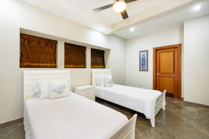 Bright room with two white single beds, wooden door, and ceiling fan.
