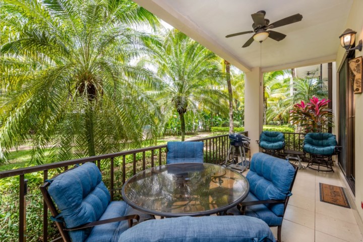 Outdoor patio with blue cushioned chairs, glass table, ceiling fan, and tropical plants.