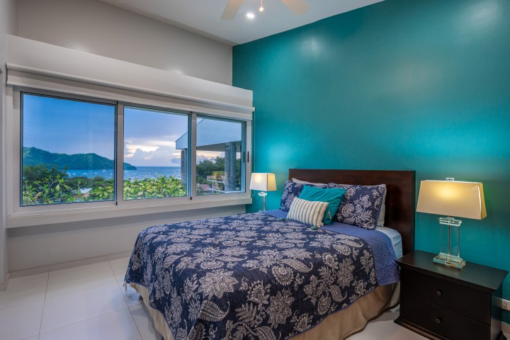 Bedroom with blue walls, patterned bedspread, and large window showing ocean view.