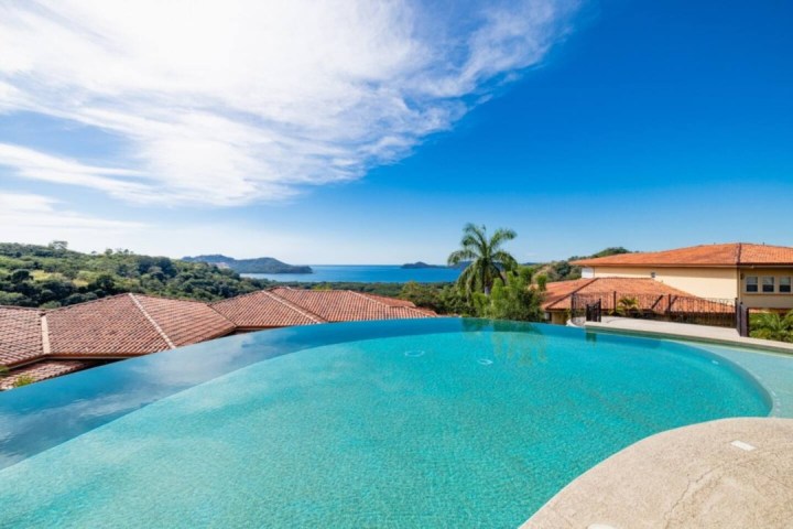 Infinity pool overlooking ocean, red-tiled roofs, and lush green landscape under blue sky.