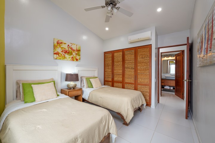 Room with two single beds, colorful pillows, wooden closet, and a bathroom entrance.