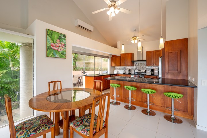 Modern kitchen with wooden cabinets, bar stools, and dining area beside large windows and wall art.