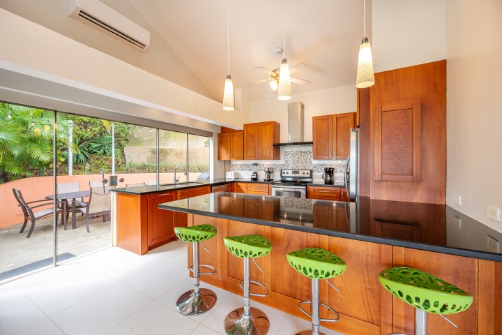 Modern kitchen with wooden cabinets, green bar stools, and large windows opening to a patio with plants.