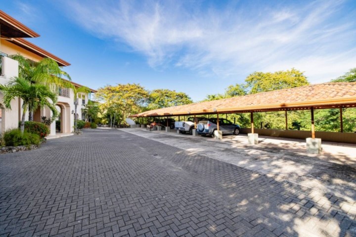 Paved driveway leads to carport with several parked cars, surrounded by greenery and buildings.