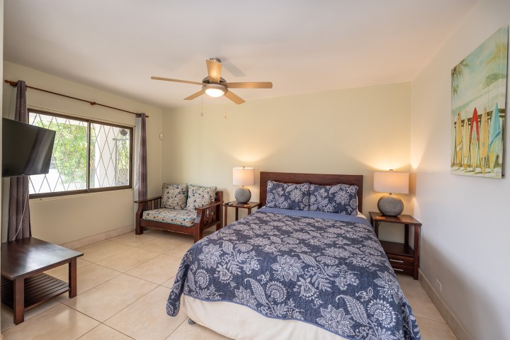 Bedroom with bed, floral bedding, nightstands, ceiling fan, armchair, window, and surfboard painting.